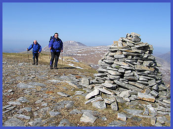 Andrew and Barry stride up to the summit cairn of Meall Horn, with Foinavon glowing in the sun behind